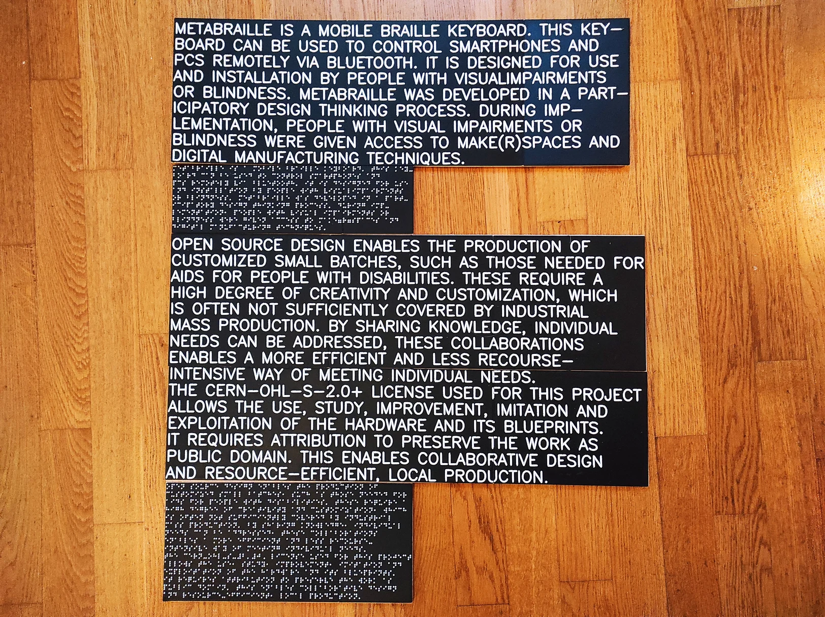 3D printed descriptions for an exhibition. The lettering is white on a black background. The text is presented in Braille and tactile print. The signs lie on a parquet floor