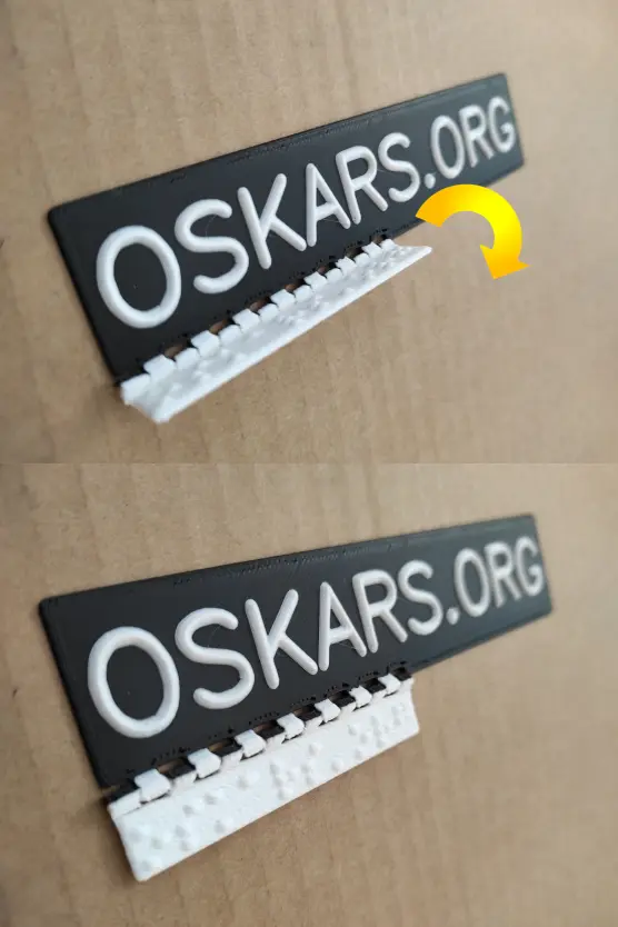 Ein Schild aus dem 3D-Drucker mit weißer Profilschrift auf schwarzem Grund. Über ein Scharnier rechtwinkelig verbunden ist ein weißer 3D-Druckteil mit Braillepunkten. Rechts neben dem Scharnier befindet sich ein gelber Pfeil, der eine Drehrichtung nach rechts darstellt. Darunter befindet dasselbe Schild mit aufgeklapptem, ebenem Brailleschriftabschnitt. Auf dem Schild steht oskars.org in fühlbaren Blockbuchstaben. Das Schild liegt auf Karton.