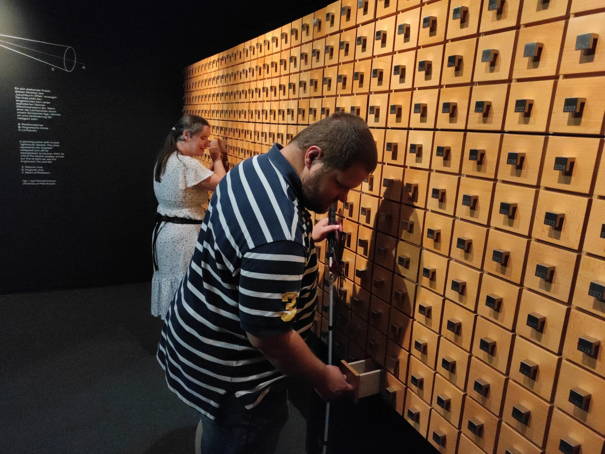 Zwei Personen vor einer Wand voll Handteller großen Schubladen aus Holz mit Metallgriffen. Der Mann im Vordergrund hält einen Blindenstock in der Linken und hält den Metallgriff einer geöffneten Schublade mit der Rechten. Er hat kurzes braunes Haar und träg einen Bart. Die Frau im Hintergrund hat ebenfalls ihre rechte Hand an einer geöffneten Schublade. Sie hat langes braunes Haar und trägt ein weißes Kleid. Die Wand im Hintergrund trägt einen Text in weißer Schrift. Die Wand und der Boden sind schwarz bzw. dunkelgrau.