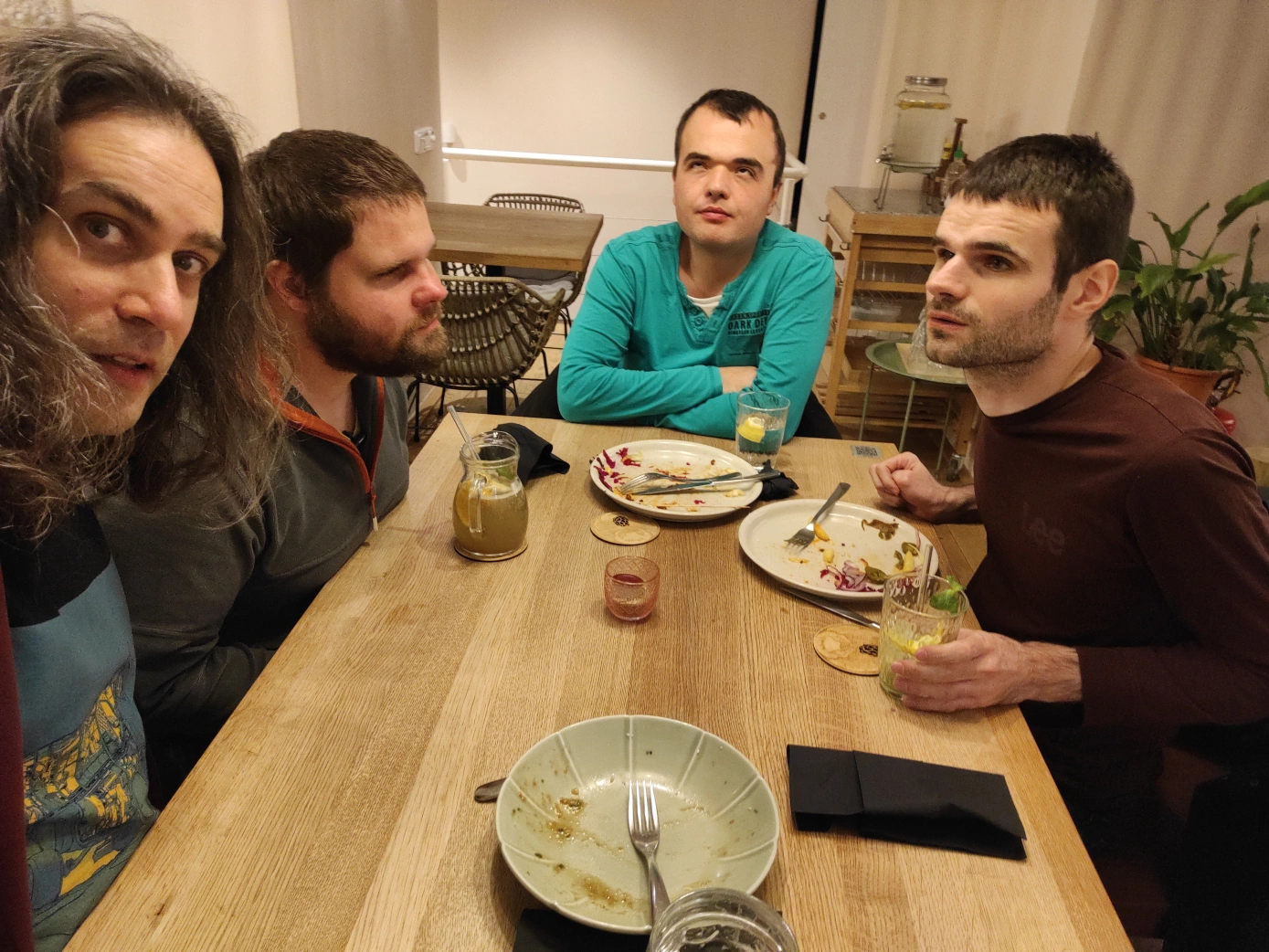 Gruppenbild von unserem Restaurantbesuch in Brünn. Vier Männer sitzen am Tisch bei bereits geleerten Tellern und noch gefüllten Gläsern. Der Tisch und auch ein Tisch im Hintergrund sowie ein Beistelltisch mit Wasserspender sind aus Holz. Die Wände sind Weiß. Eine Topfpflanze steht im Hintergrund.