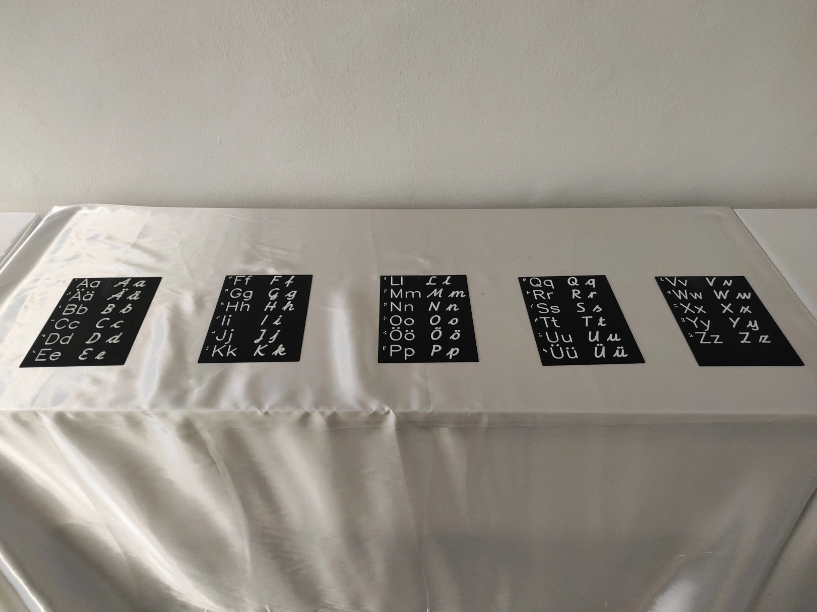 3D-gedruckte Alphabete, Braille-Alphabet, taktile Druckbuchstaben und Schreibschrift in Groß- und Kleinbuchstaben. Die Zeichen sind weiß und die Grundplatten sind schwarz. Fünf Tafeln liegen auf einem Tisch mit weißem Tischtuch vor einer weißen Wand.